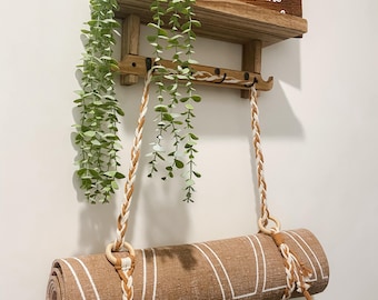 May include: A rolled-up brown yoga mat with white stripes hangs from a wooden shelf with a braided rope and wooden rings. The shelf also holds a small sign that reads "Drink Wine" and a trailing green plant.