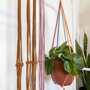 May include: A brown terracotta pot filled with green leafy plants hangs from a white metal stand. The plant is suspended by a colorful macrame plant hanger with strands of brown, pink, blue, and yellow cords. Additional macrame hangers are visible.