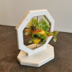 Pode incluir: Suporte octogonal de madeira branca com um pequeno espeto de salada dentro. O espeto de salada é feito de alface, cenouras e fruta amarela.