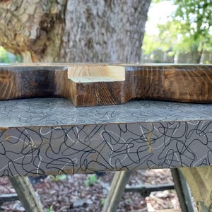 Puede incluir: Un bloque de madera grueso y rectangular con un acabado te&ntilde;ido en marr&oacute;n oscuro y una muesca central de color claro sin pintar en uno de los lados. La veta de la madera es visible en toda la superficie oscura, mostrando texturas naturales.