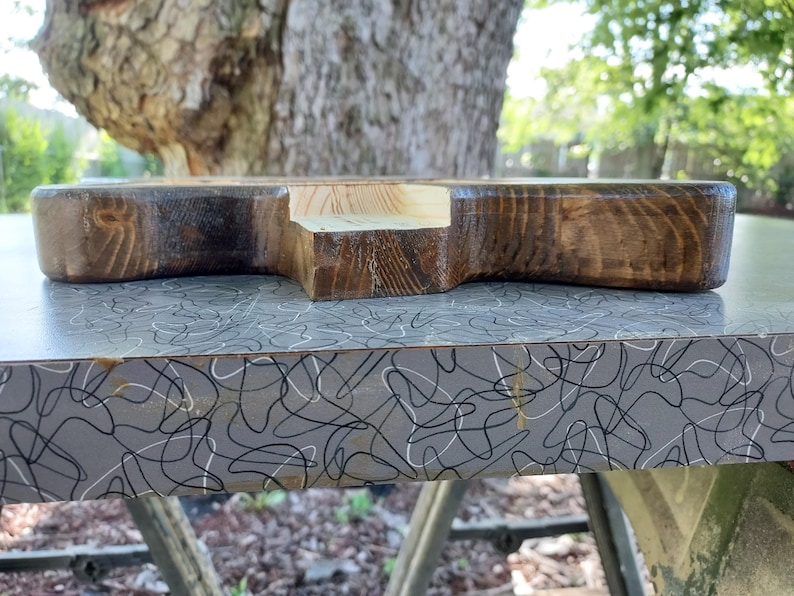 Puede incluir: Un bloque de madera grueso y rectangular con un acabado te&ntilde;ido en marr&oacute;n oscuro y una muesca central de color claro sin pintar en uno de los lados. La veta de la madera es visible en toda la superficie oscura, mostrando texturas naturales.