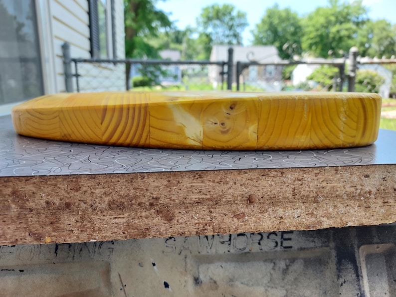 Pu&ograve; includere: Un tagliere rotondo in legno, di colore giallo chiaro. &Egrave; realizzato con pi&ugrave; pezzi di legno, che mostrano il motivo della venatura. Il tagliere &egrave; appoggiato su una superficie a motivi. Diametro: circa 30 cm.