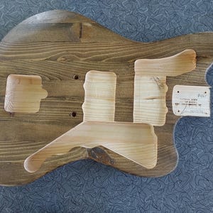 Puede incluir: Un cuerpo de guitarra el&eacute;ctrica de madera sin terminar con un acabado te&ntilde;ido en marr&oacute;n oscuro. El cuerpo presenta cavidades fresadas para la electr&oacute;nica y el hardware, con una etiqueta rectangular blanca adherida al lado derecho con texto escrito a mano.