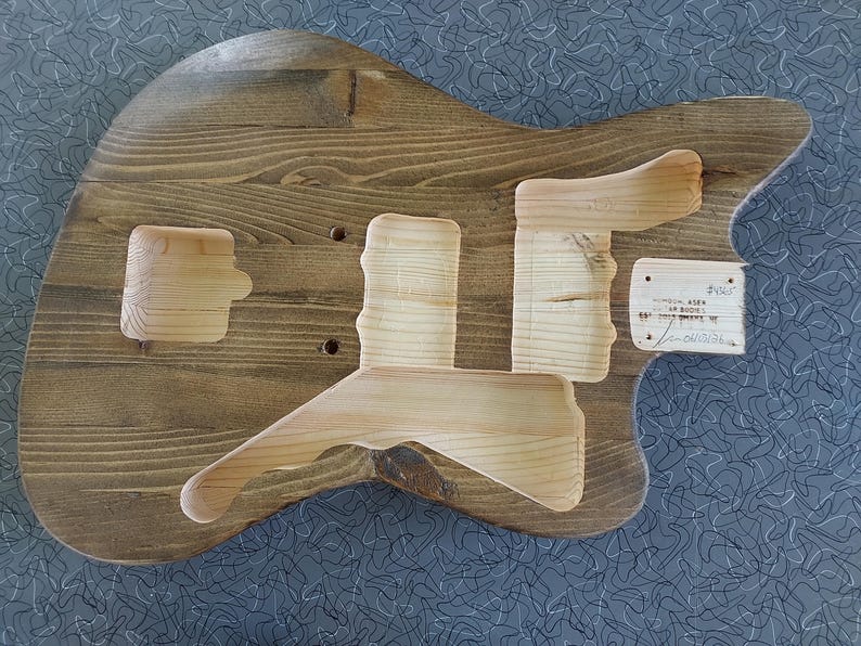 Puede incluir: Un cuerpo de guitarra el&eacute;ctrica de madera sin terminar con un acabado te&ntilde;ido en marr&oacute;n oscuro. El cuerpo presenta cavidades fresadas para la electr&oacute;nica y el hardware, con una etiqueta rectangular blanca adherida al lado derecho con texto escrito a mano.
