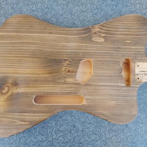 May include: A stained wooden guitar body with cutouts for pickups, controls, and neck. The wood grain is visible, and the body has a natural, rustic appearance. A small wooden plate with text is attached near the neck pocket.