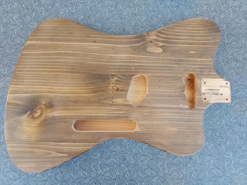 May include: A stained wooden guitar body with cutouts for pickups, controls, and neck. The wood grain is visible, and the body has a natural, rustic appearance. A small wooden plate with text is attached near the neck pocket.