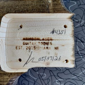 Op de afbeelding: Close-up van een houten gitaarbody-stuk met de tekst "#4351", "NAMOONLASER GUITAR BODIES EST. 2015 MAMA NE", en de datum "05/07/26" erop geschreven. Het hout heeft een natuurlijke nerf en is omgeven door een grijs patroon.