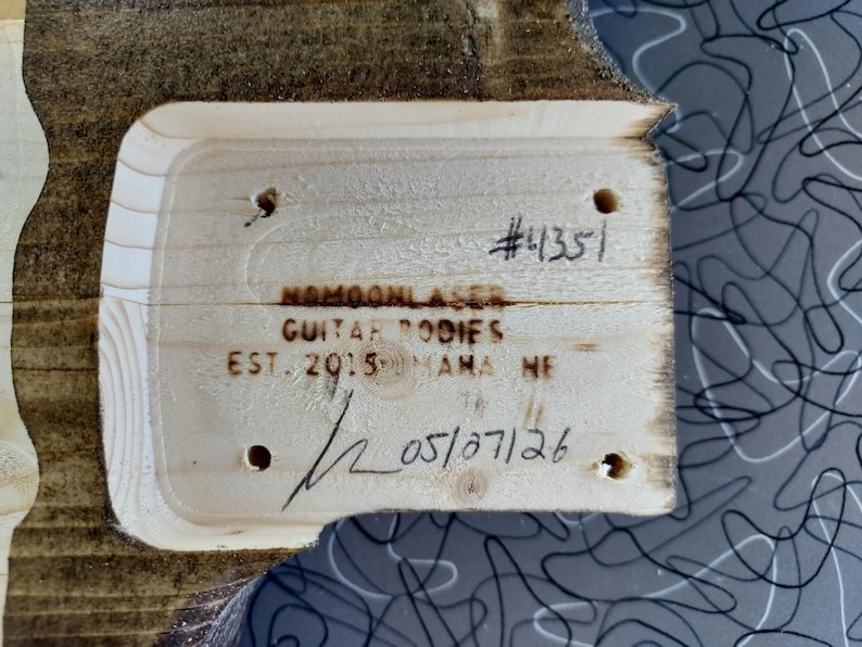 Op de afbeelding: Close-up van een houten gitaarbody-stuk met de tekst "#4351", "NAMOONLASER GUITAR BODIES EST. 2015 MAMA NE", en de datum "05/07/26" erop geschreven. Het hout heeft een natuurlijke nerf en is omgeven door een grijs patroon.