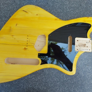 Pu&ograve; includere: Un corpo di chitarra in legno giallo con battipenna nero e ritagli per pickup e controlli. Il corpo ha una venatura naturale del legno e una finitura liscia. Una piccola etichetta di legno &egrave; attaccata con testo.