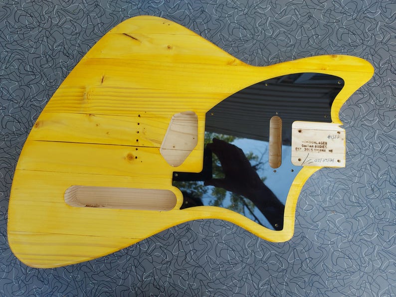 Pu&ograve; includere: Un corpo di chitarra in legno giallo con battipenna nero e ritagli per pickup e controlli. Il corpo ha una venatura naturale del legno e una finitura liscia. Una piccola etichetta di legno &egrave; attaccata con testo.