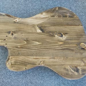 Op de afbeelding: Een gitaarvormige houten snijplank met een natuurlijke houtnerf afwerking. De plank heeft een glad oppervlak en afgeronde randen, met een lichtbruine kleur. De houtnerf is zichtbaar.