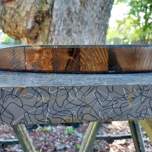 Puede incluir: Una tabla de cortar circular de madera con un acabado te&ntilde;ido oscuro y patrones de veta de madera visibles. El borde de la tabla muestra una construcci&oacute;n segmentada con tonos marrones variados, desde el bronceado claro hasta tonos caf&eacute; oscuro.