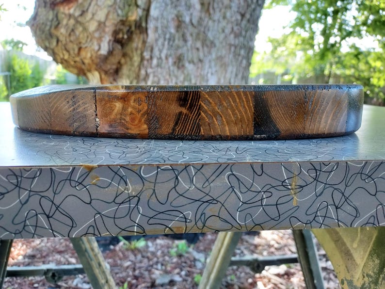 Puede incluir: Una tabla de cortar circular de madera con un acabado te&ntilde;ido oscuro y patrones de veta de madera visibles. El borde de la tabla muestra una construcci&oacute;n segmentada con tonos marrones variados, desde el bronceado claro hasta tonos caf&eacute; oscuro.