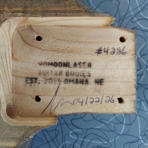 May include: Close-up of a wooden guitar body with carved edges. The wood has a light brown color and visible grain. Text includes "#4386", "40MOONLASER GUITAR BODIES EST. 2015 OMAHA NE" and "04/22/26".