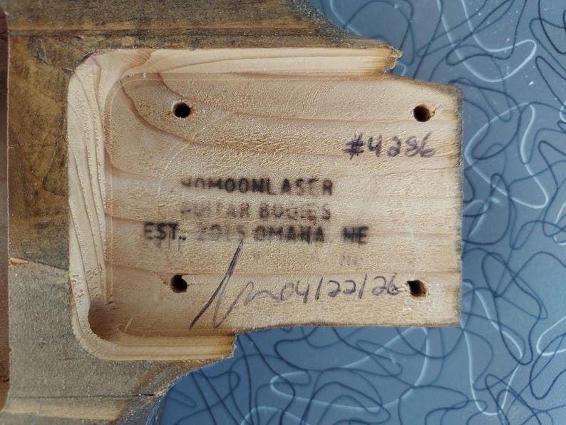 May include: Close-up of a wooden guitar body with carved edges. The wood has a light brown color and visible grain. Text includes "#4386", "40MOONLASER GUITAR BODIES EST. 2015 OMAHA NE" and "04/22/26".