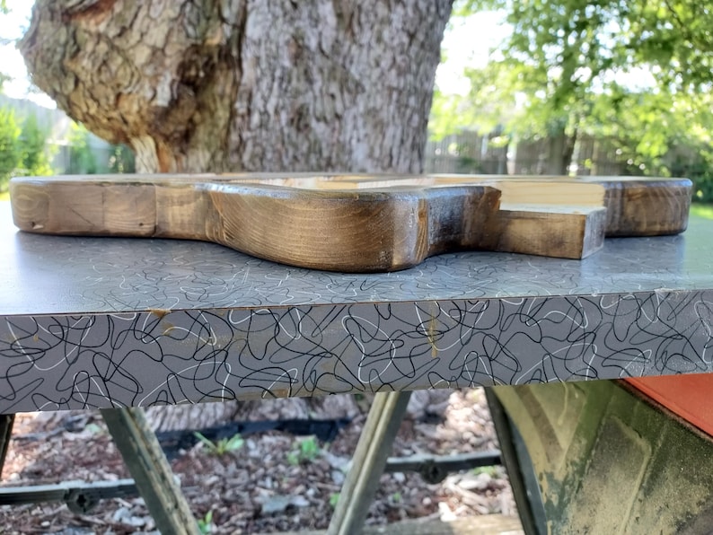 Puede incluir: Una tabla de madera gruesa y sin terminar con un borde oscuro, te&ntilde;ido y texturizado. La pieza de madera presenta un patr&oacute;n de grano natural y una superficie superior lisa y clara, mostrando el material crudo y la artesan&iacute;a del objeto.