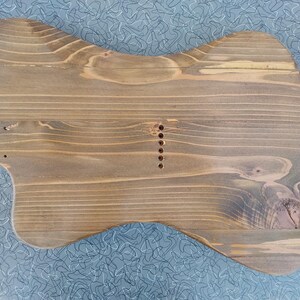 May include: A wooden guitar body, unfinished, with a natural wood grain. The body is shaped like a classic electric guitar, with a contoured design and pre-drilled holes for components. The wood has a warm, brown tone with visible grain patterns.