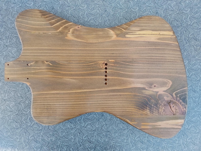May include: A wooden guitar body, unfinished, with a natural wood grain. The body is shaped like a classic electric guitar, with a contoured design and pre-drilled holes for components. The wood has a warm, brown tone with visible grain patterns.