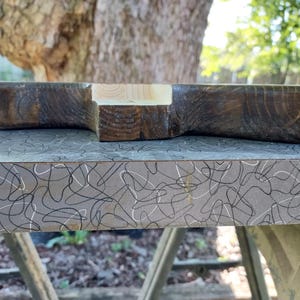 Op de afbeelding: Een handgemaakt houten serveerplank met een donkerbruine afwerking en een accent van natuurlijk hout. De plank heeft een unieke vorm en wordt gepresenteerd op een patroonoppervlak, wat de rustieke charme benadrukt.