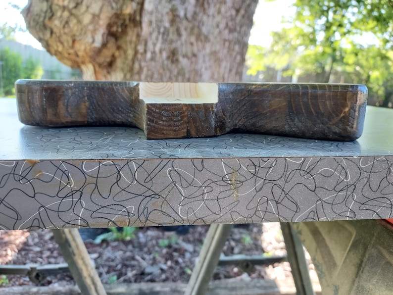 Op de afbeelding: Een handgemaakt houten serveerplank met een donkerbruine afwerking en een accent van natuurlijk hout. De plank heeft een unieke vorm en wordt gepresenteerd op een patroonoppervlak, wat de rustieke charme benadrukt.