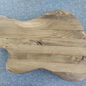 Puede incluir: Un cuerpo de guitarra en bruto construido con varias tablas horizontales de madera marr&oacute;n claro con vetas y nudos visibles. La pieza presenta una forma asim&eacute;trica contorneada con un recorte en el lado izquierdo y una superficie lisa sin acabado.