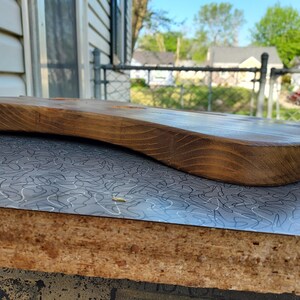 May include: A handcrafted, wooden guitar body with a natural finish. The body is curved and has a smooth, polished surface. The wood grain is visible, and the color is a warm brown. The guitar body is resting on a gray and white patterned surface.