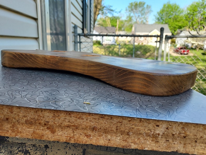 May include: A handcrafted, wooden guitar body with a natural finish. The body is curved and has a smooth, polished surface. The wood grain is visible, and the color is a warm brown. The guitar body is resting on a gray and white patterned surface.