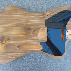 May include: A wooden guitar body with a natural finish. The body has cutouts for pickups, a neck, and other components. A blue pickguard is visible, reflecting the sky. A small wooden piece with text is attached.