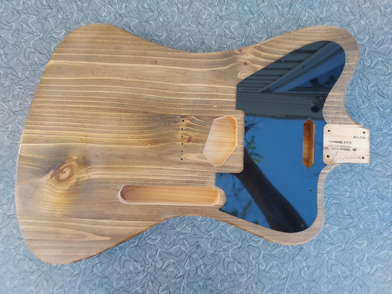 May include: A wooden guitar body with a natural finish. The body has cutouts for pickups, a neck, and other components. A blue pickguard is visible, reflecting the sky. A small wooden piece with text is attached.