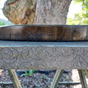 Op de afbeelding: Een ronde, donkerbruine houten snijplank met een zichtbaar houtnerfpatroon. Het bord is gemaakt van verschillende stukken hout die aan elkaar zijn gezet. Het bord ligt op een grijs oppervlak met een zwart-wit krulpatroon.