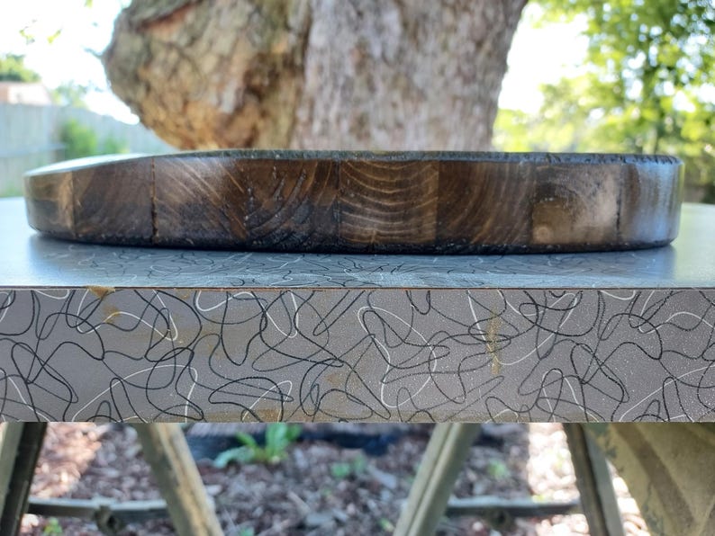 Op de afbeelding: Een ronde, donkerbruine houten snijplank met een zichtbaar houtnerfpatroon. Het bord is gemaakt van verschillende stukken hout die aan elkaar zijn gezet. Het bord ligt op een grijs oppervlak met een zwart-wit krulpatroon.