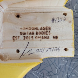 Pu&ograve; includere: Primo piano di un corpo di chitarra in legno con il testo "-MOONLASER GUITAR BODIES EST. 2015 OMAHA NE" e il numero "#4352". &Egrave; visibile anche la data "05/27/26".
