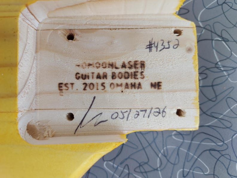 Pu&ograve; includere: Primo piano di un corpo di chitarra in legno con il testo "-MOONLASER GUITAR BODIES EST. 2015 OMAHA NE" e il numero "#4352". &Egrave; visibile anche la data "05/27/26".