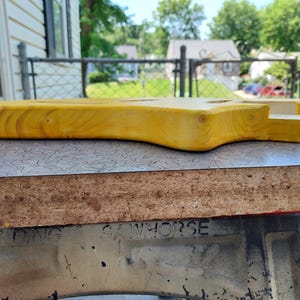 Pu&ograve; includere: Un corpo di chitarra incompiuto in legno giallo chiaro. Il corpo &egrave; stato levigato e poggia su una superficie a motivi. Il corpo della chitarra &egrave; in fase di costruzione. Il corpo della chitarra &egrave; lungo circa 76 cm.