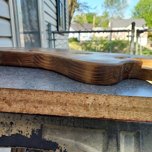 May include: A partially constructed, unfinished wooden guitar body. The wood has a dark brown stain with visible grain patterns. The body rests on a gray and silver patterned surface, with a glimpse of a sawhorse in the foreground.