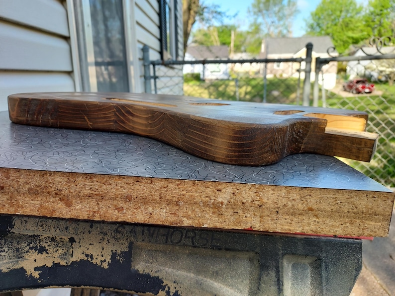 May include: A partially constructed, unfinished wooden guitar body. The wood has a dark brown stain with visible grain patterns. The body rests on a gray and silver patterned surface, with a glimpse of a sawhorse in the foreground.