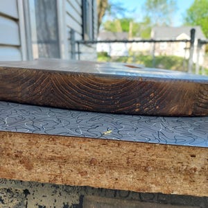 May include: A thick, oval-shaped wooden cutting board with a rich, dark brown color. The wood grain is prominently displayed, and the board has a smooth, rounded edge. The board is resting on a surface with a gray and white pattern.