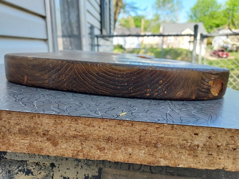 May include: A thick, oval-shaped wooden cutting board with a rich, dark brown color. The wood grain is prominently displayed, and the board has a smooth, rounded edge. The board is resting on a surface with a gray and white pattern.