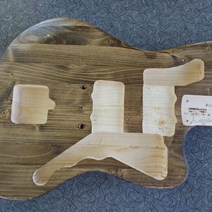 Op de afbeelding: Een houten gitaarbody met een donkerbruine beits. Er zijn uitsparingen voor de pickups, brug en hals. Lichte houten stukken zijn bevestigd. Een klein label met tekst is bevestigd.
