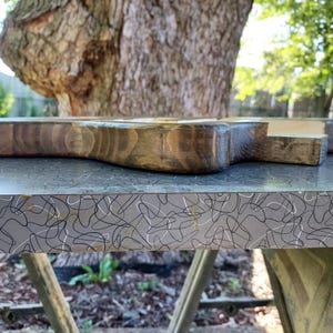 Op de afbeelding: Een houten snijplank met een donkere vlek, met een handvat en een rechthoekige vorm. De plank ligt op een grijze ondergrond met een wit en zwart abstract patroon. De achtergrond toont een boom en groen.