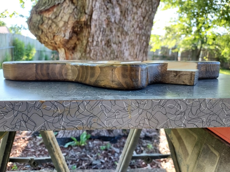 Op de afbeelding: Een houten snijplank met een donkere vlek, met een handvat en een rechthoekige vorm. De plank ligt op een grijze ondergrond met een wit en zwart abstract patroon. De achtergrond toont een boom en groen.