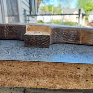 May include: A handcrafted wooden cutting board with a dark brown finish. The board has a unique shape with a handle-like extension. The wood grain is visible, adding to its rustic charm. The board is placed on a surface with a blue and white pattern.