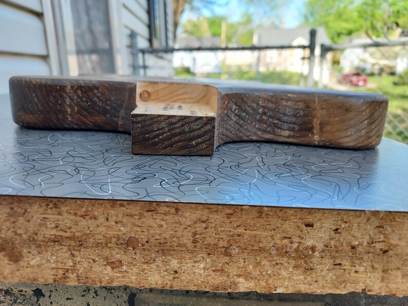 May include: A handcrafted wooden cutting board with a dark brown finish. The board has a unique shape with a handle-like extension. The wood grain is visible, adding to its rustic charm. The board is placed on a surface with a blue and white pattern.