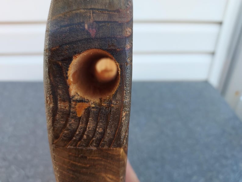 May include: Close-up of a section of dark brown wood with a drilled hole. The hole is cylindrical and appears to be freshly drilled. The wood grain is visible, and the edges of the hole show some splintering.