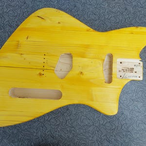 Pu&ograve; includere: Un corpo di chitarra giallo in legno, con ritagli per pickup e controlli. Il corpo ha una finitura liscia e un motivo naturale delle venature del legno. Una piccola etichetta di legno &egrave; attaccata con testo.