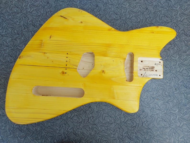 Pu&ograve; includere: Un corpo di chitarra giallo in legno, con ritagli per pickup e controlli. Il corpo ha una finitura liscia e un motivo naturale delle venature del legno. Una piccola etichetta di legno &egrave; attaccata con testo.