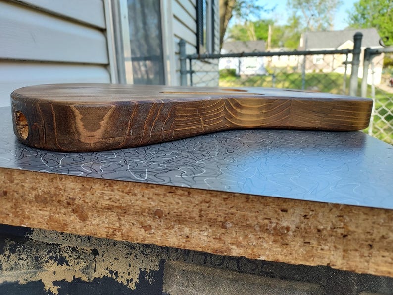 May include: A handcrafted, wooden guitar body with a natural, stained finish. The body is shaped with smooth curves and a hole for the neck. The wood grain is visible, showcasing the natural texture and color variations. The guitar body is resting on a patterned surface.