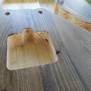 Puede incluir: Un cuerpo de guitarra el&eacute;ctrica de madera sin terminar que presenta un interior de madera clara natural dentro de las cavidades de las pastillas fresadas y un acabado te&ntilde;ido en gris oscuro desgastado en la superficie exterior.