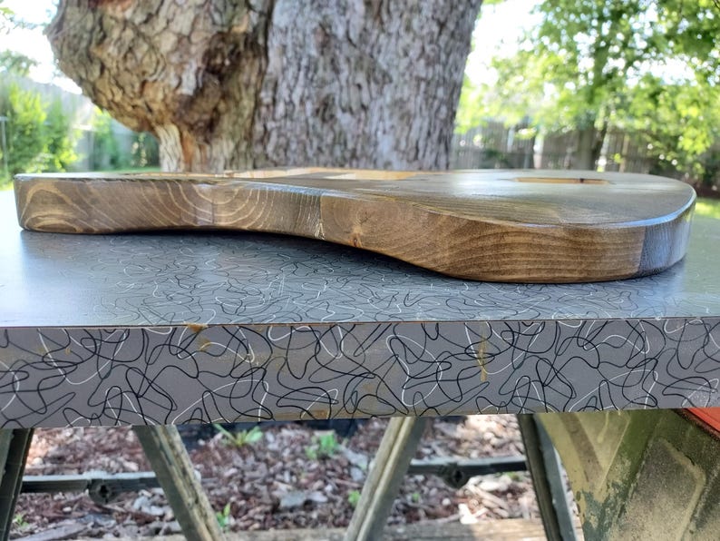 Puede incluir: Un cuerpo de guitarra el&eacute;ctrica de madera maciza con un acabado liso te&ntilde;ido de marr&oacute;n oscuro que muestra patrones naturales de veta de madera. El cuerpo presenta un borde contorneado y m&uacute;ltiples cavidades fresadas para componentes.