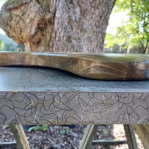 Op de afbeelding: Een houten snijplank in donkerbruin met een handvat. De plank heeft een gladde, gebogen vorm en ligt op een grijze ondergrond met een wit en zwart wervelpatroon. Op de achtergrond zijn een boom en groen te zien.